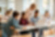 Three middle school students sitting at a desk with their notebooks and watching a laptop. A teacher is standing behind them and using the laptop mouse pad.