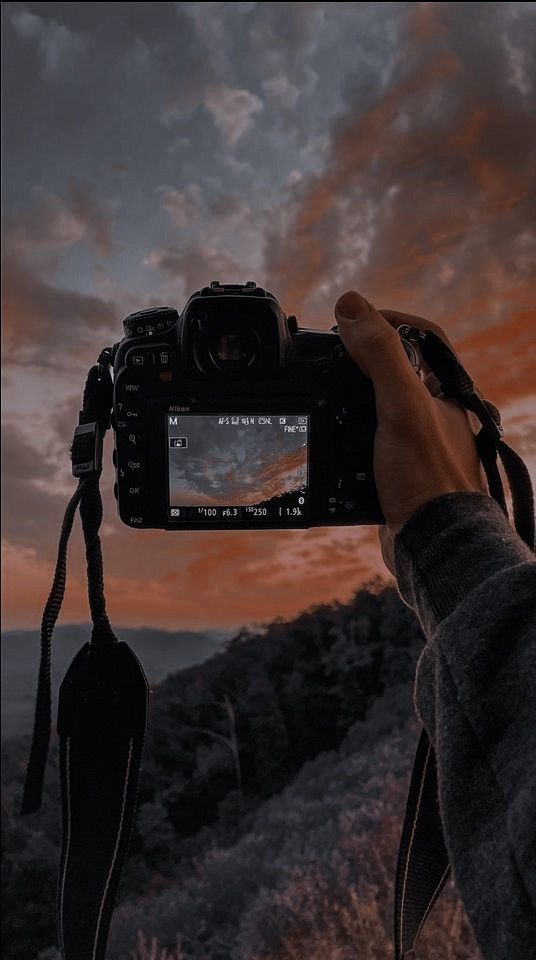 Hand holding a camera against a dramatic sunset sky, showing the same scene on the screen. Forested hills in the background. Warm tones.