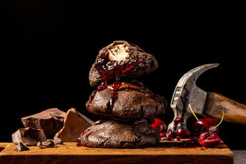 A stock of chocolate cookies on a cutting board with a hammer next to crushed cherries and chunks of chocolate