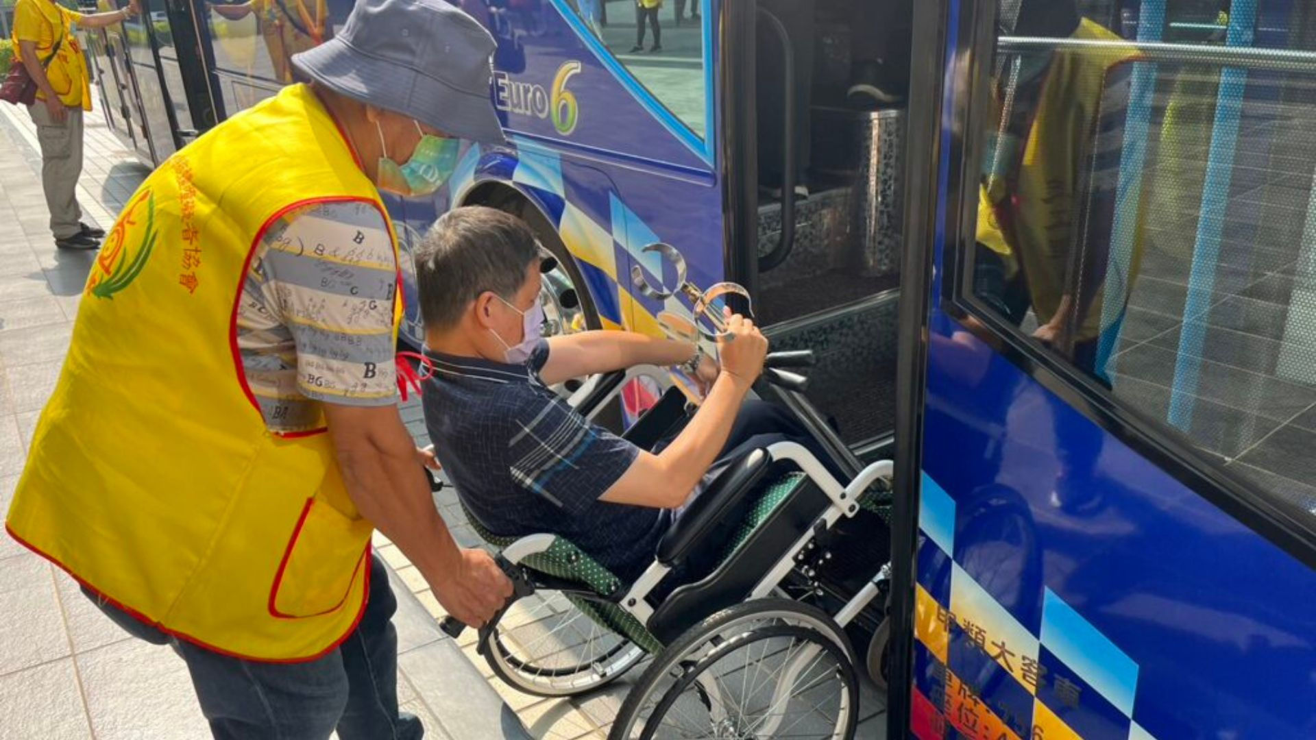 一位身穿黃背心志工正協助坐輪椅的會員搭乘無障礙遊覽車，雙方表情認真，背景為亮眼藍色車身。