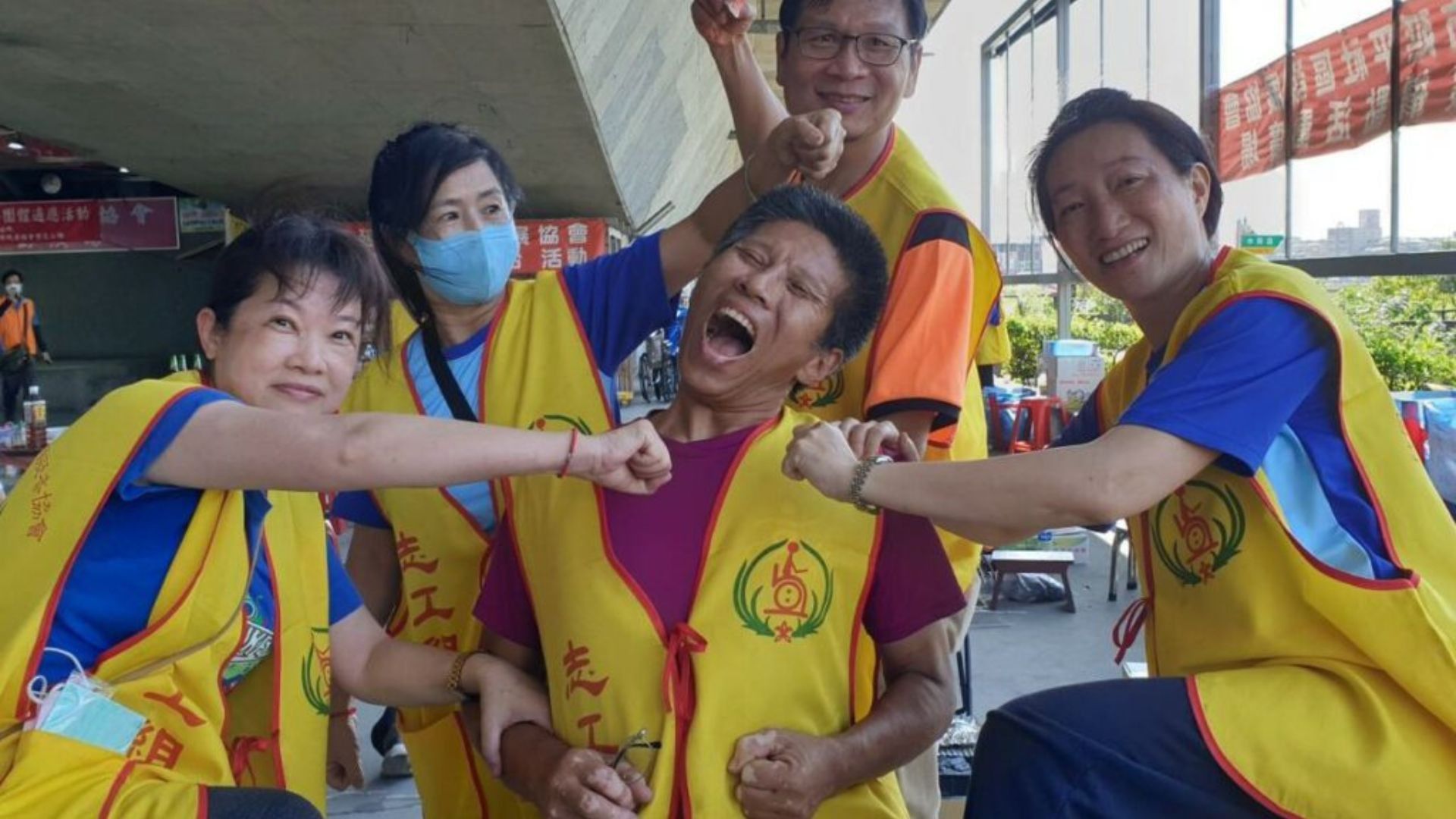 穿著黃色志工背心的夥伴在戶外開心互動，其中一人做出搞笑表情，其他人環繞模仿出拳，氣氛輕鬆愉快。