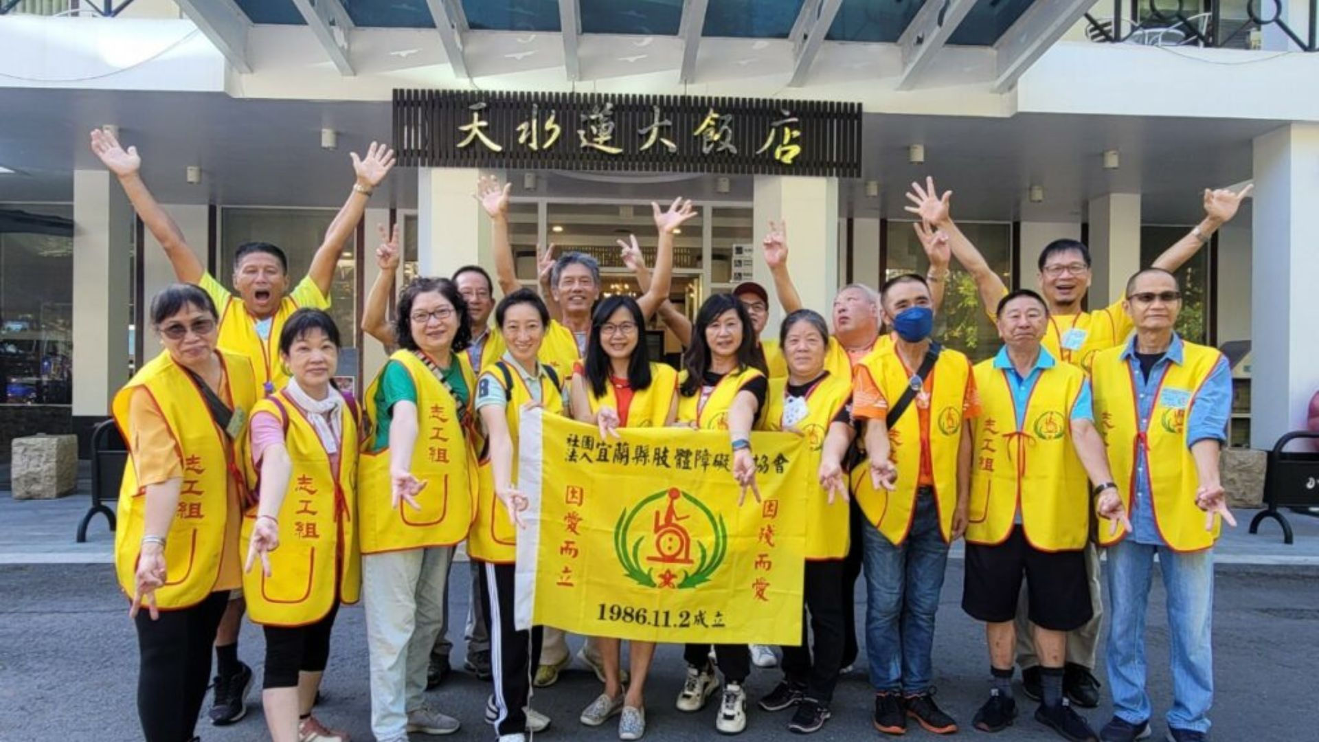 全體志工於大飯店門前高舉雙手合影，黃背心上「志工組」字樣醒目，展現團隊凝聚力與行動力。