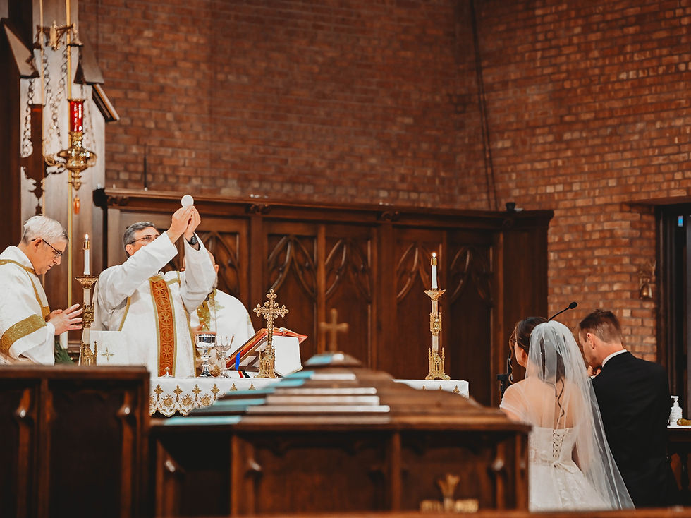 Priest in white robe lifts the Eucharist in a church, with a couple kneeling nearby. Brick walls, wooden pews, and candles set a solemn mood.
