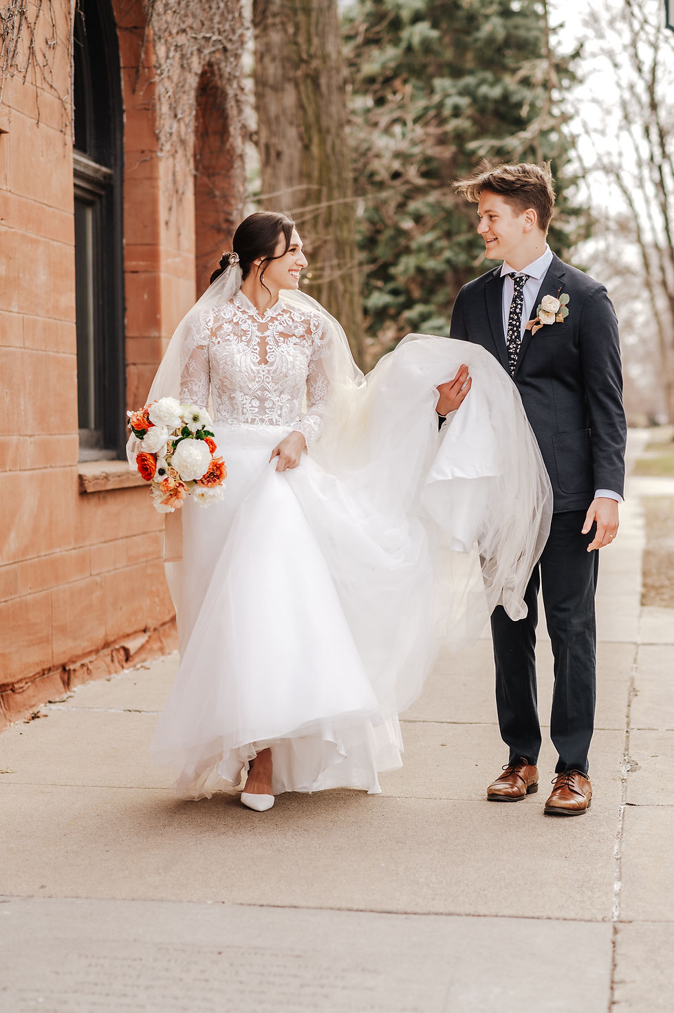 Bride in lace dress holds bouquet, groom in suit holds her dress. They walk on sidewalk, smiling. Background: brick wall, trees.