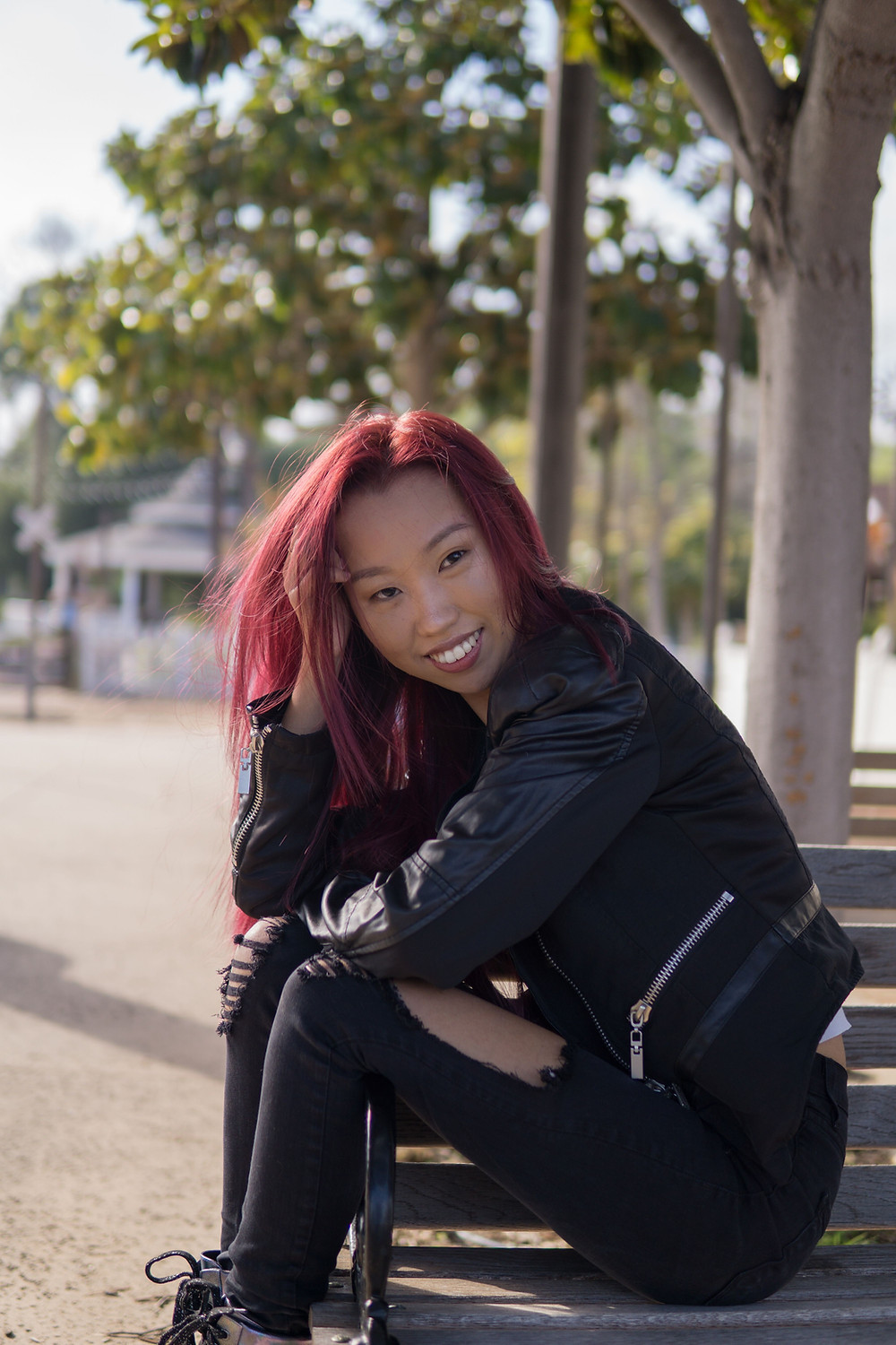A picture of me, a young woman, sitting on a park bench sideways, with my legs resting over the arm rest. My hand is in my long red hair while I rest my head on it.