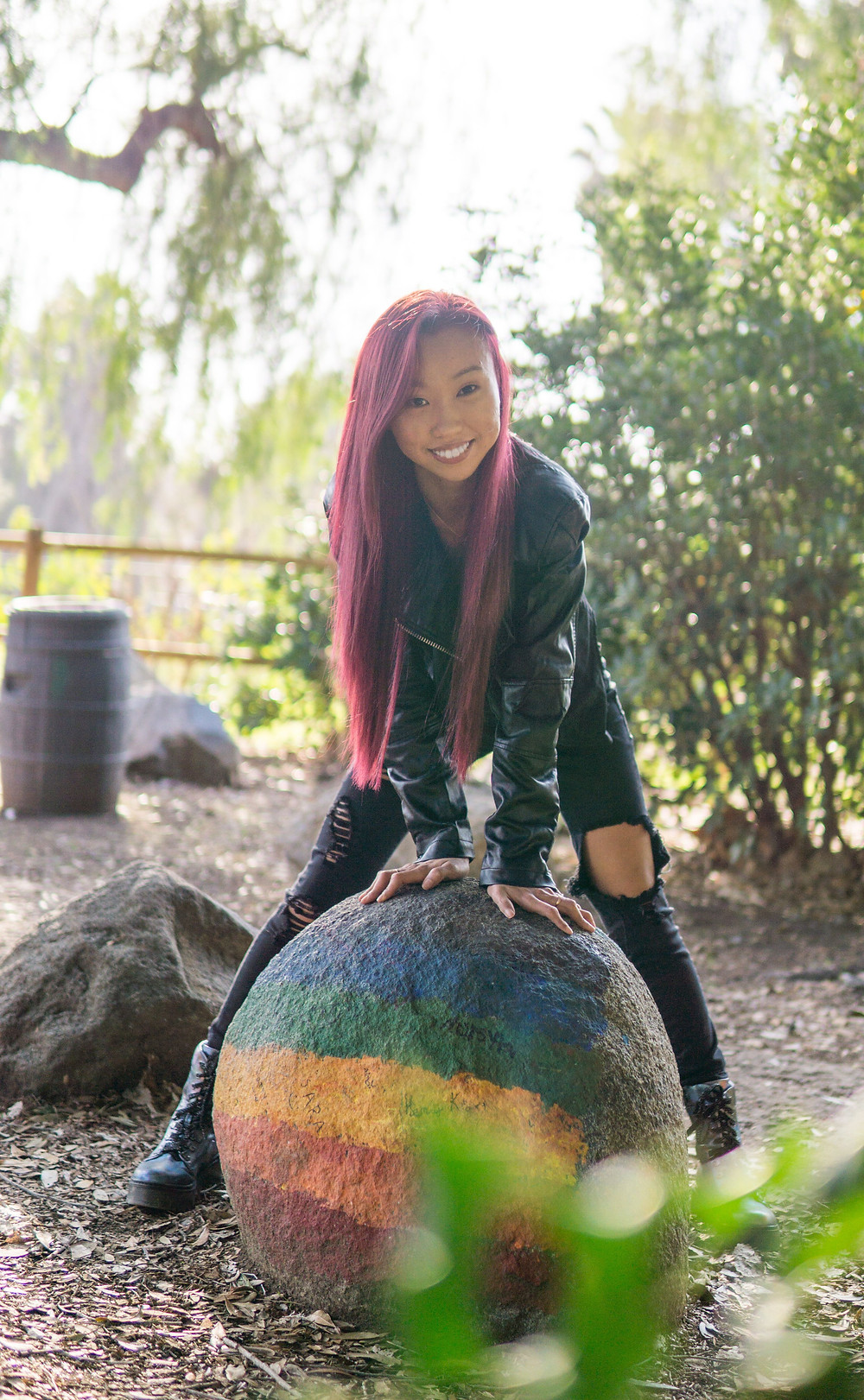I, a young woman, with long dyed-red hair, am wearing a black leather jacket and black torn denim pants. I am hunched over and resting both hands on a rainbow-painted boulder while smiling.
