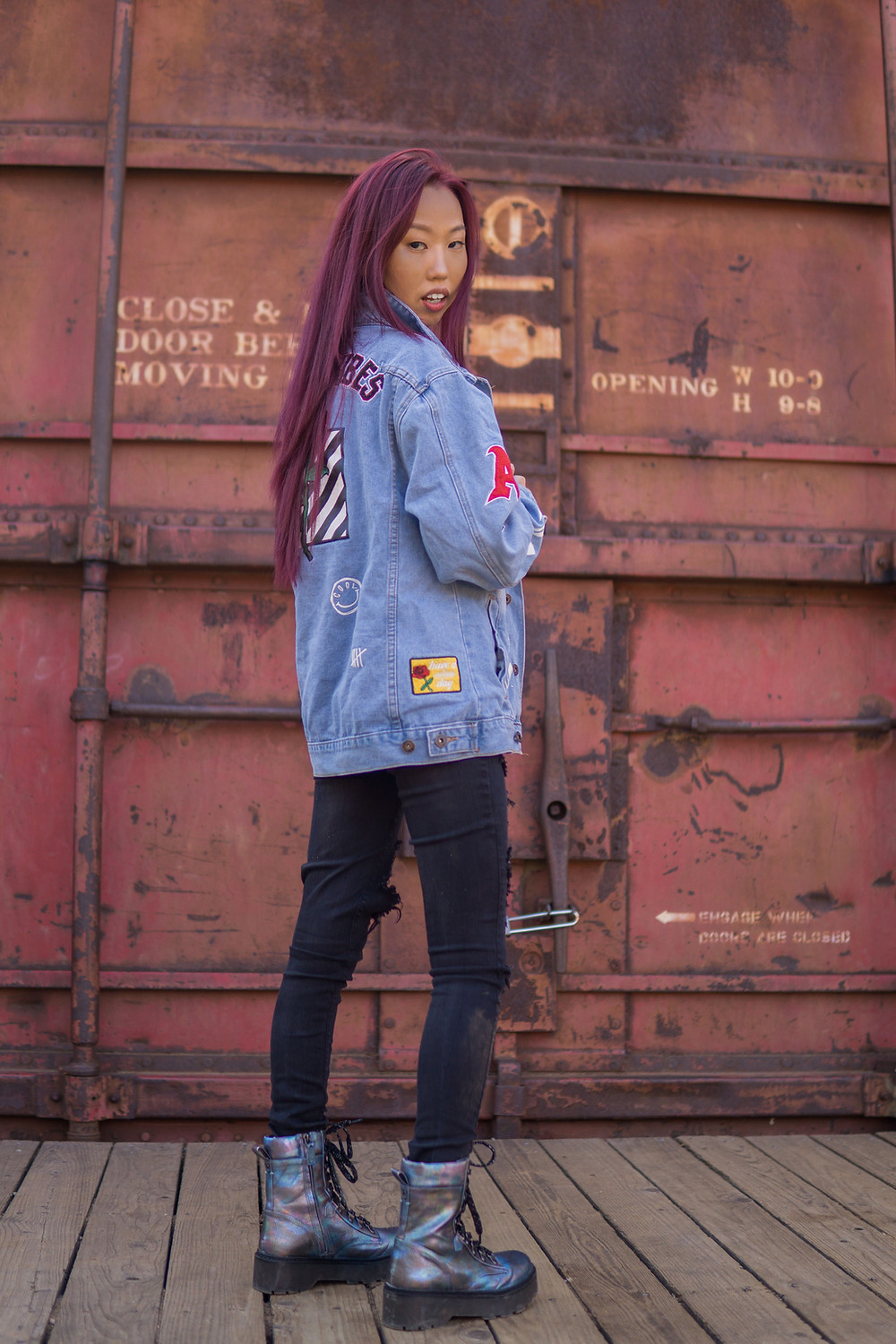 I, a young woman, wearing black torn denim pants, iridescent combat boots and a light blue denim jacket with various patches sewn on it, am standing with my body facing away from the camera toward rust-colored shipping crates in the background, but my face turns and looks toward the camera. I have a neutral expression.