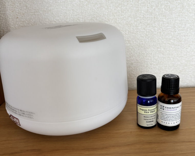 木製テーブルに白いアロマディフューザーと、ティーツリーとラベンダーの精油瓶2本が並ぶ静かな室内の写真