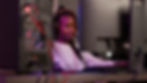 A young Black woman with headphones on looking at a computer in the foreground.