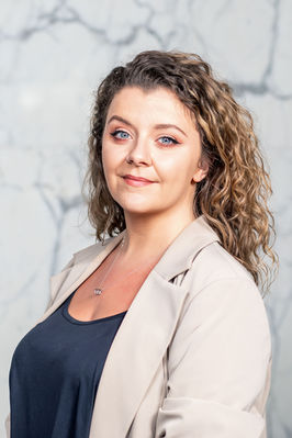 Corporate portrait of woman with curly hair and nose ring, beige blazer, marble background.