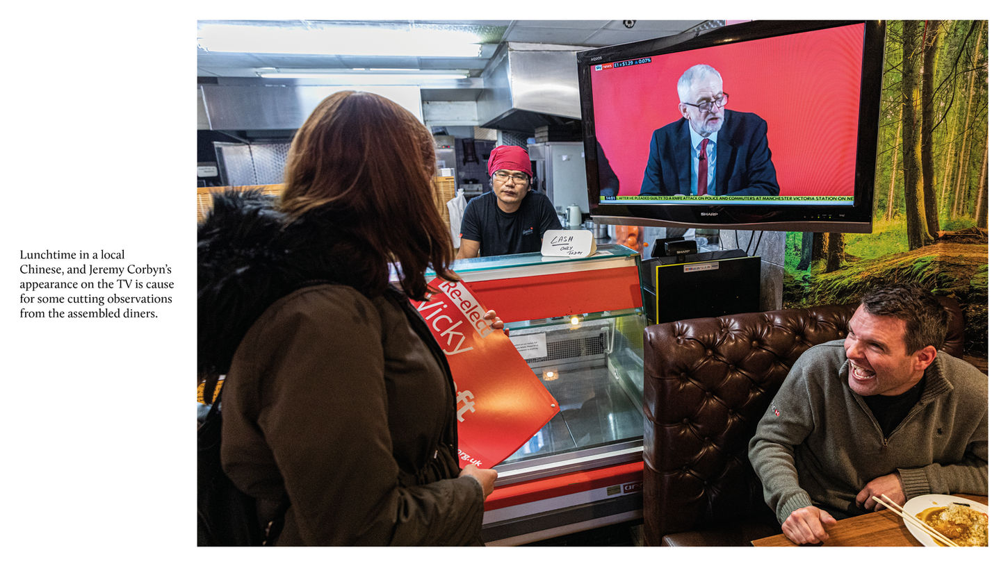 In a café, a television shows a political speech while a diner laughs and a campaigner holds a red poster at the counter.