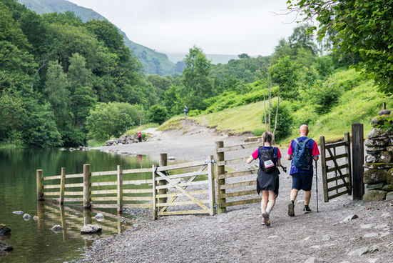 Two Cancer Research UK Big Hike participants walk beside a lake through green Lake District countryside.