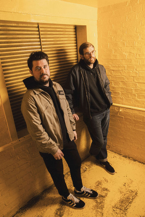 Two men in casual jackets stand in a narrow yellow stairwell, looking to camera.