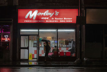 Night street scene of a Morley’s fried chicken takeaway in London; worker behind the counter, glowing red signs, and people walking past on a rainy pavement.