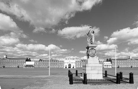 Wide black and white photo of a formal parade ground and long brick barracks building, dominated by a central stone statue beneath a sky of billowing clouds.