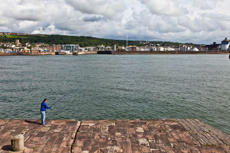 Person fishing from a stone harbour wall with town and marina across the water.