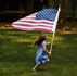 Barefoot girl in a blue dress runs across a grassy yard, carrying a large American flag on a pole.