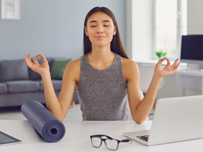 A young woman practicing mindfulness meditation at her desk, sitting in a relaxed posture with eyes closed and hands in a mudra gesture, next to a yoga mat, laptop, tablet, and glasses in a calm home office setting.