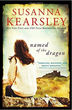 Book cover of Susanna Kearsley’s named of the dragon, showing a woman in a sweater sitting on rocks in a windswept field near ruins.