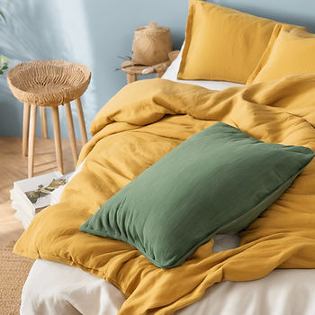 A beautifully arranged bedroom with a mustard yellow duvet and green pillow on a bed, next to a natural wooden stool and light blue walls.