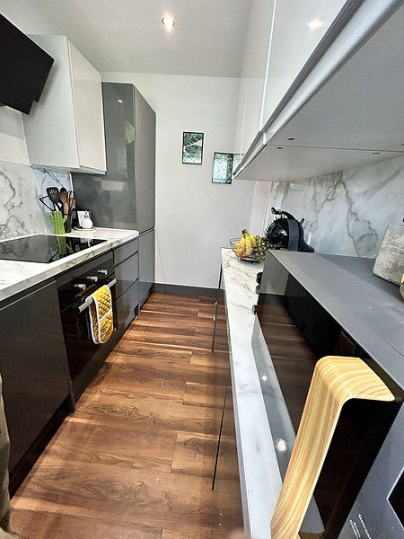 Sleek, narrow kitchen with dark grey cabinets, white countertops, and wood floor.