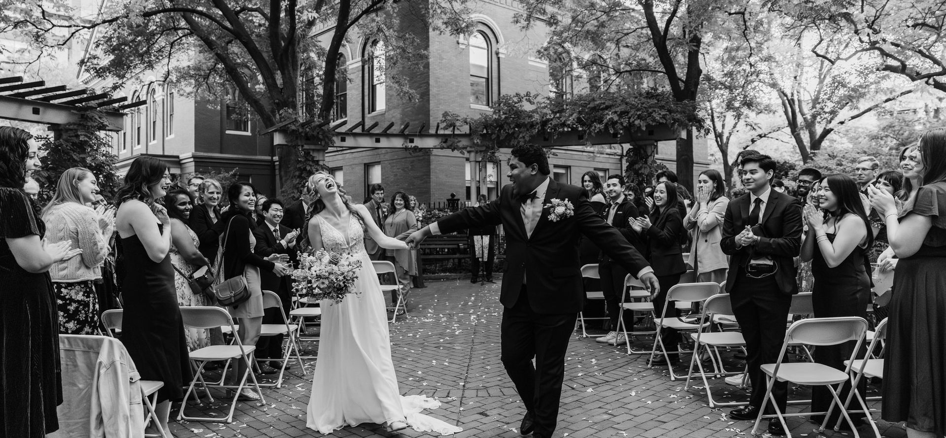 Black and white image of a smiling couple walking down the aisle after their wedding, with guests cheering