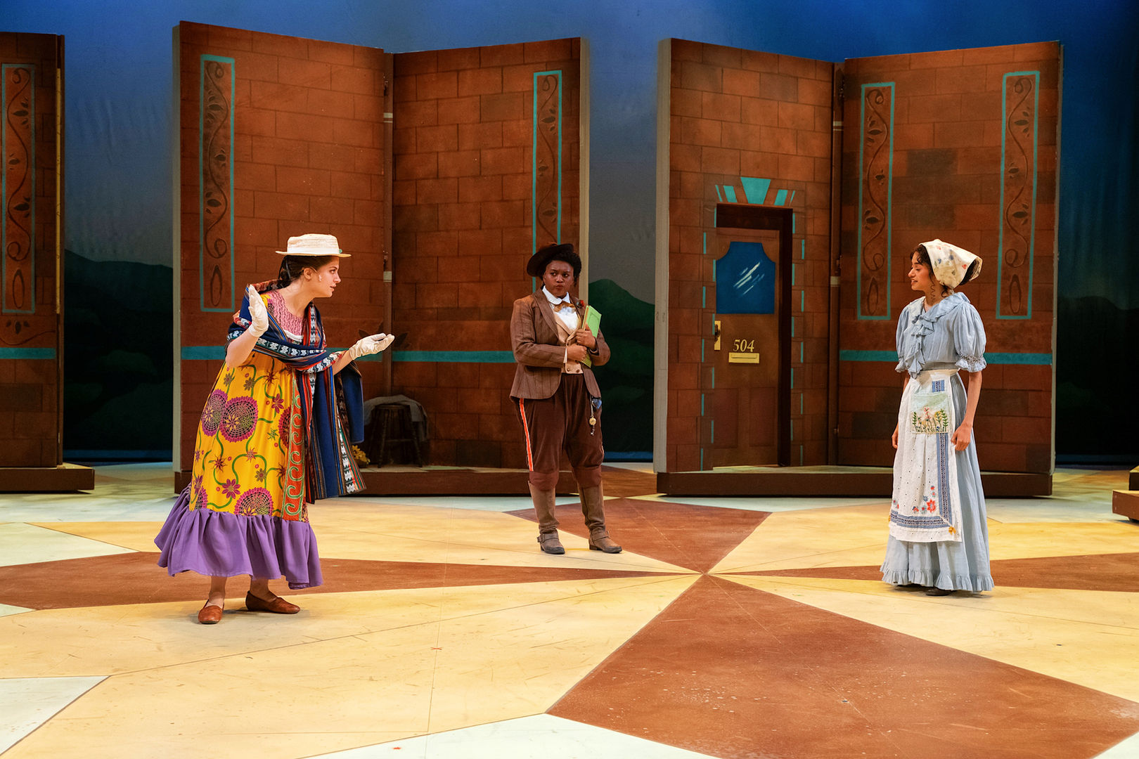 Three actors in period costumes perform on a geometric theatrical stage.