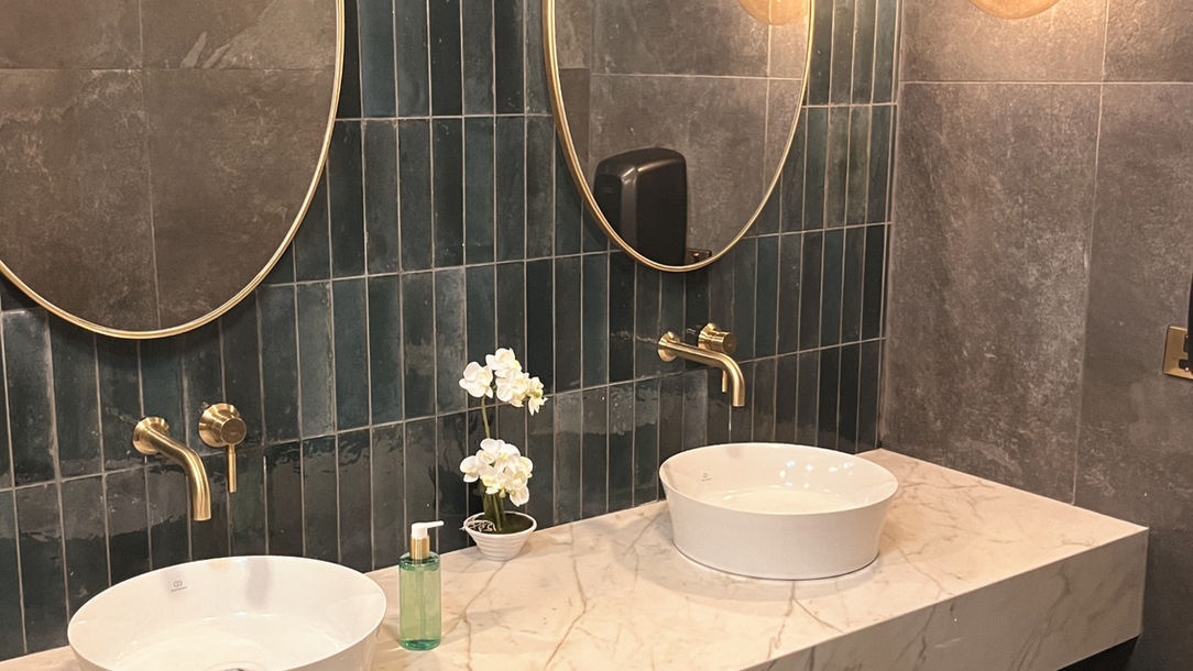 Modern bathroom interior design by MOIRÉ Architects in Nicosia featuring a floating marble vanity with two white vessel sinks, wall-mounted brass faucets, oval mirrors against a vertical emerald-green tile wall, and round brass sconces illuminating a dark-stone floor and walls.