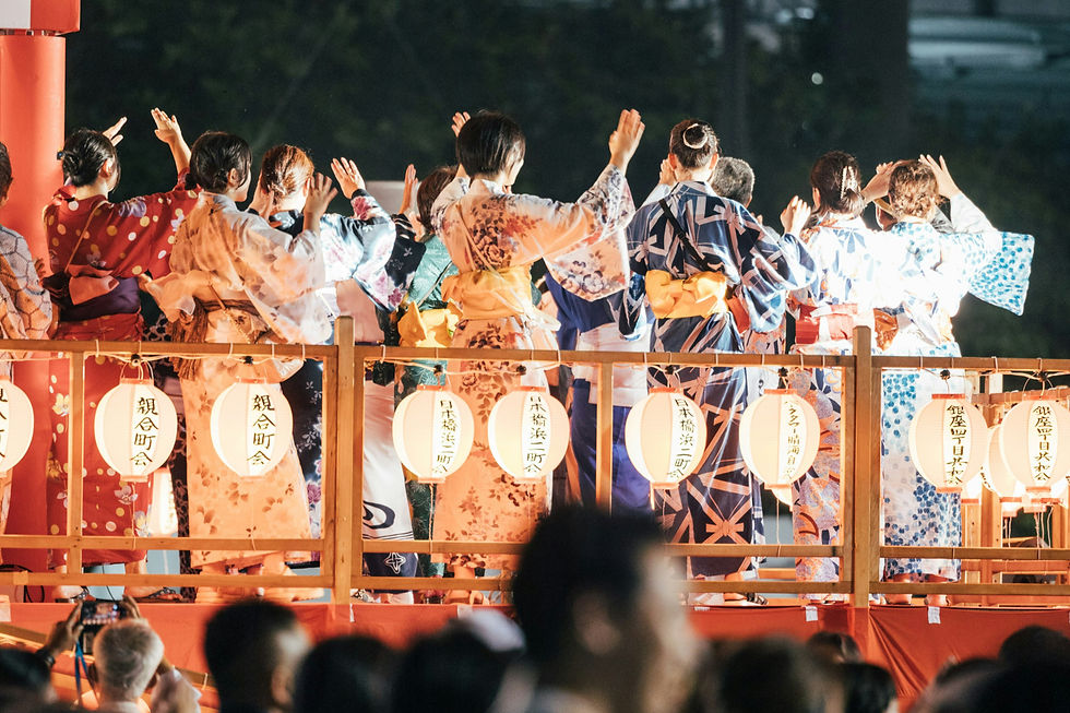 Women in colorful yukata raise their hands in a nighttime Japanese festival, with glowing lanterns and a festive crowd.