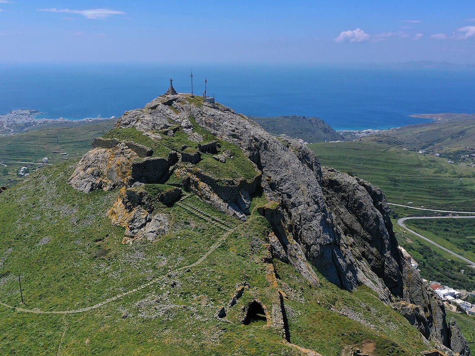 Rocky hilltop with crosses overlooks blue ocean and green fields below. Path winds through vibrant landscape under clear blue sky.