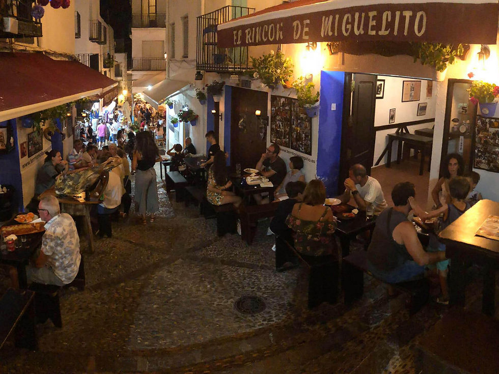 People dine outdoors at night on a cobblestone street. Restaurant name "El Rincon de Miguelito" displayed. Warm, lively setting.