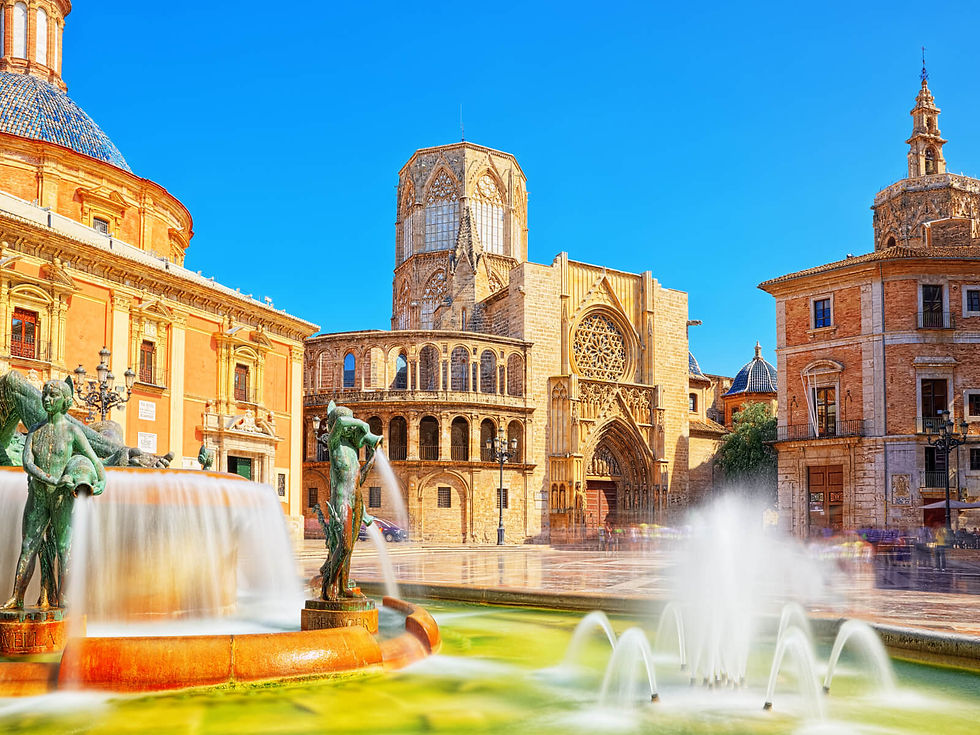 Historic plaza with a central fountain and bronze statues in front of a cathedral. Clear blue sky and vibrant architecture create a lively mood.