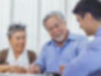A smiling elderly couple with grey hair sits at a table across from a younger man in a blue shirt, presumably an advisor or agent, discussing documents.