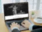 Hands typing on a laptop displaying a website with an image of people dining. Background shows a bright room with a plant and coffee cup.