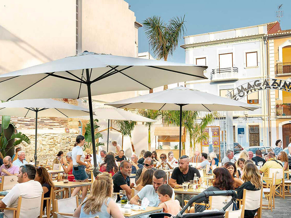 People dining outdoors under umbrellas at a bustling café. The setting is sunny with palm trees.