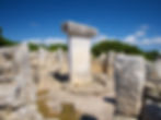 Ancient stone ruins with tall rectangular pillars under a clear blue sky. Green bushes border the site, conveying a sense of history.