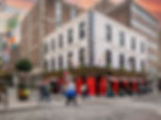 Street view of The Auld Dubliner pub with a colorful entrance and red accents. People walk by on cobblestone streets. Irish flag visible.