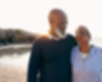 A happy, smiling elderly Black couple walking on a beach at sunset. The man has his arm around the woman, who is wearing a blue and white striped shirt and necklace.