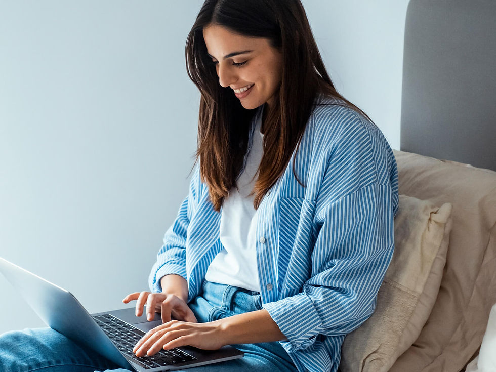 A woman in a blue striped shirt smiles while typing on a laptop, sitting on a bed with cream pillows in a softly lit room.
