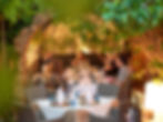 People dining outdoors at a table under greenery, surrounded by stone arches. Warm lighting, glasses, and plates on the table. Cozy atmosphere.