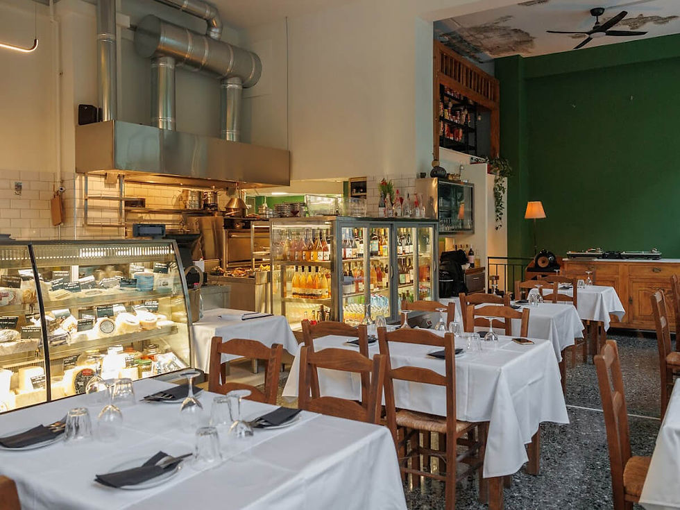 Cozy restaurant with wooden chairs, white tablecloths, and a green accent wall. Displays of cheese and drinks; warm, inviting atmosphere.