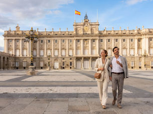 A couple in light suits walks arm in arm in front of the Royal Palace of Madrid, with a Spanish flag flying above. Clear blue sky. Header image related to the blog post "Battle over the new world of exclusivity: Madrid, Athens and the paradox of the high-spend traveller