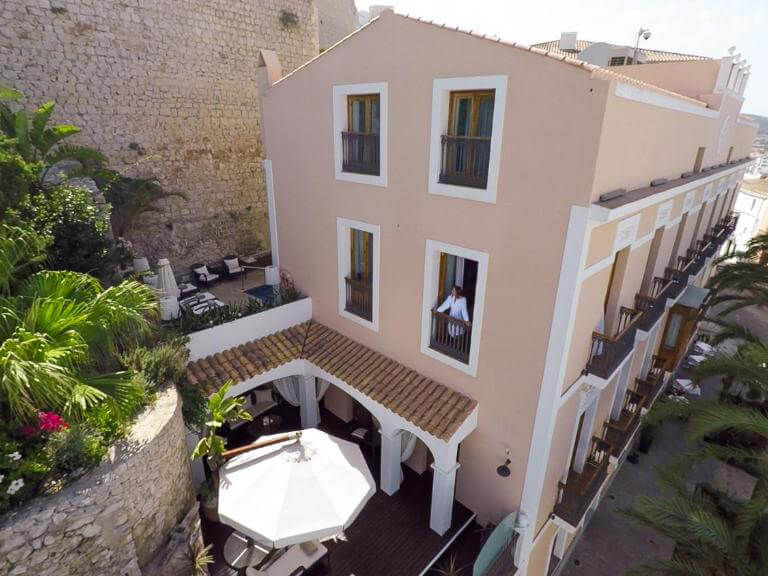 Pink building with white trim, large windows, and a person looking out. Outdoor patio with plants and umbrellas. Stone wall backdrop.