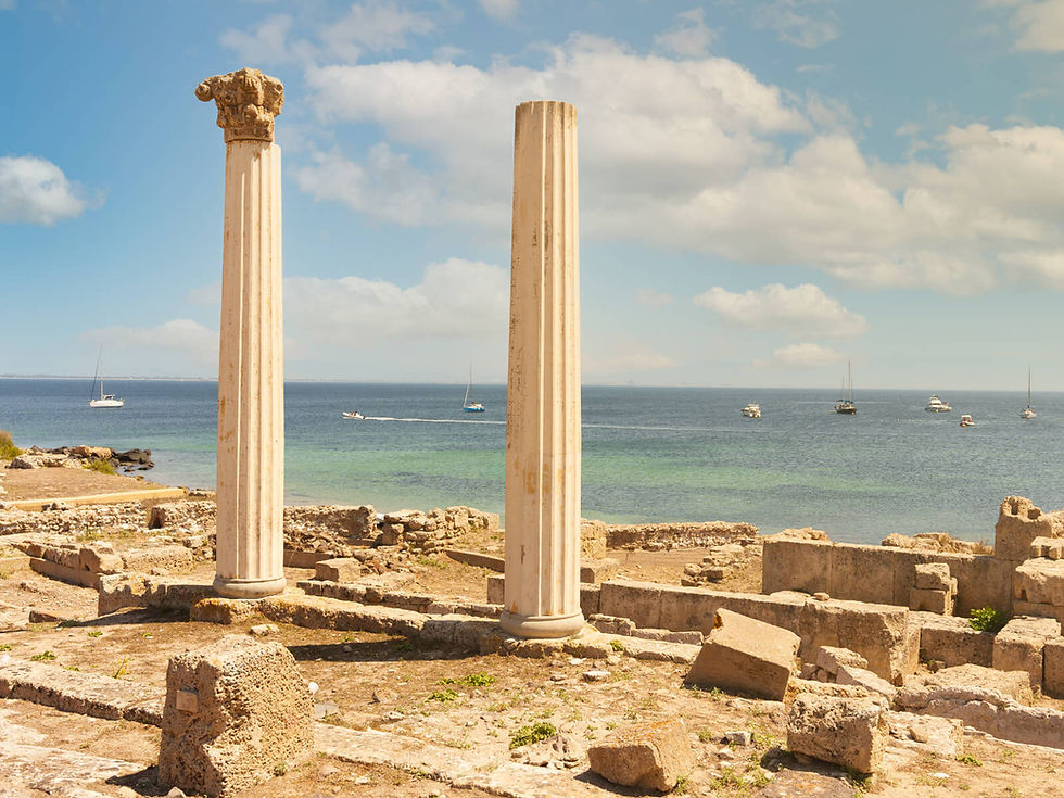 Two ancient columns stand among ruins by the sea. Sailboats are visible on the blue horizon under a partly cloudy sky, creating a serene atmosphere.