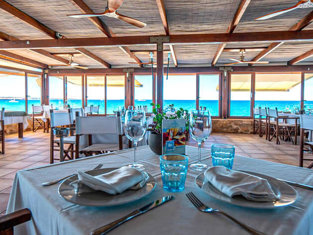 Table for two with wine glasses and blue water view in seaside restaurant, wooden ceiling, fans, and vibrant outdoor light creating a serene mood.