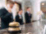 Hotel reception desk with a brass bell in focus. Three blurred staff in suits talk on phones in the background, creating a busy, formal mood.