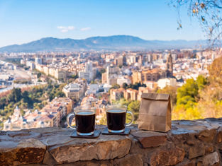 Two coffee mugs and a paper bag on a stone ledge, overlooking Málaga. Header image related to the blog post "Specialty Coffee in Málaga"
