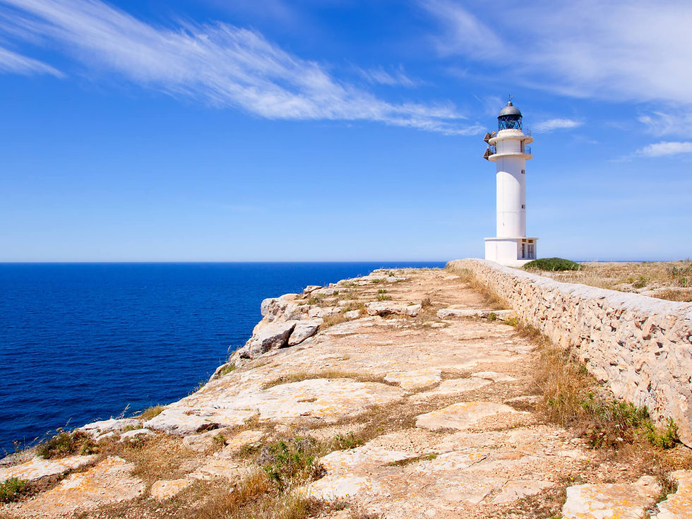White lighthouse on a rocky coastal cliff, under a bright blue sky with wispy clouds. The calm sea stretches into the distance.