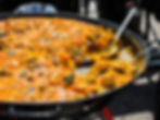 A large pan of vibrant yellow paella with seafood like mussels and shrimp. A serving spoon rests inside. Outdoor setting, sunlight.