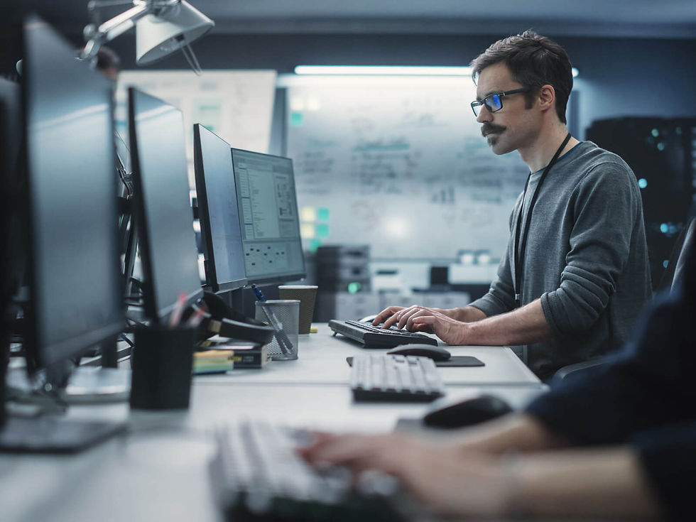 Hombre con gafas trabaja en computadora en oficina moderna. Monitores negros y pizarra blanca de fondo. Ambiente profesional y concentrado.
